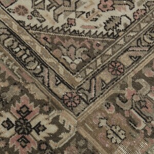 May include: Close-up of a vintage rug with a detailed pattern. The rug features a combination of beige, brown, and pink tones. The design includes floral and geometric elements, creating a textured and aged appearance.
