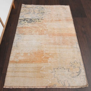 May include: A vintage-style rug with a faded, distressed look. The rug has a light beige background with a subtle pattern of faded orange and gray. The rug has a fringed edge.