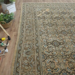 May include: An overhead shot of a large, rectangular, vintage rug with an intricate floral pattern in shades of beige, brown, and blue. The rug is placed on a wooden floor, with a wooden easel and paint bottles in the foreground.