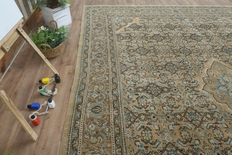 May include: An overhead shot of a large, rectangular, vintage rug with an intricate floral pattern in shades of beige, brown, and blue. The rug is placed on a wooden floor, with a wooden easel and paint bottles in the foreground.