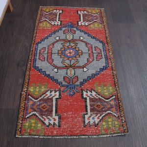 May include: A small, rectangular rug with a distressed, vintage appearance. The rug features a central gray medallion with floral and geometric patterns in orange, blue, and green against a red background. The rug's edges are bordered with similar patterns.