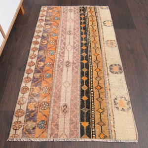 Puede incluir: Una alfombra vintage con fondo crema y patrones geométricos coloridos en tonos naranja, marrón, negro y morado. La alfombra tiene un borde con flecos.