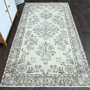 Puede incluir: Una alfombra vintage de color crema con un patrón floral en tonos marrones, beige y rosa. La alfombra tiene un borde con un diseño geométrico.