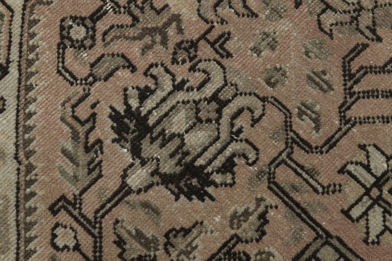 May include: Close-up of a section of a vintage rug with a detailed floral pattern. The rug features a colour palette of muted pink, beige, and black, with intricate designs and a textured surface. The rug's design is symmetrical.