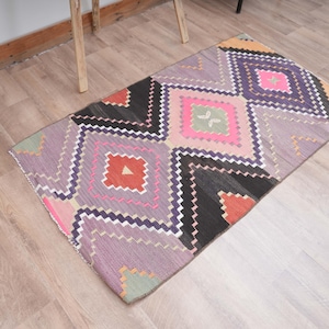 May include: A rectangular woven rug featuring a geometric pattern of diamonds and zigzags in shades of lavender, black, bright pink, orange, and light green. The design consists of repeating shapes arranged in horizontal rows across the surface.
