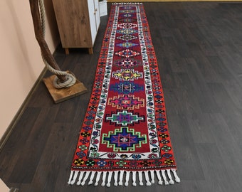 Op de afbeelding: Een lang, rood loper met een witte rand en ingewikkelde geometrische patronen in tinten rood, blauw, groen, geel en oranje. De loper heeft aan beide uiteinden witte franjes.