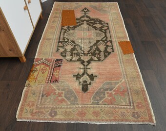Puede incluir: Una alfombra rectangular de estilo vintage con un diseño geométrico central en negro y marrón sobre un fondo rosa. La alfombra presenta parches de tela naranja y estampada, con un aspecto descolorido y desgastado. Los bordes están deshilachados.