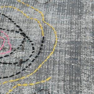 May include: A gray rug with a pink, yellow, and black embroidered design. The design features a large circle with a smaller circle inside, and a curved line.