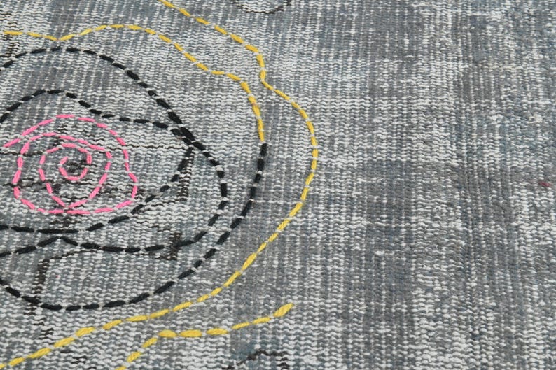 May include: A gray rug with a pink, yellow, and black embroidered design. The design features a large circle with a smaller circle inside, and a curved line.