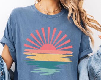 May include: Blue jean-colored t-shirt with a colorful sunset graphic. The graphic features a pink and orange sun with yellow, green, and blue horizontal stripes below, resembling a reflection. The shirt is size XL.