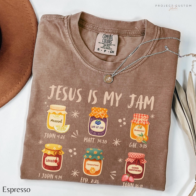 K&ouml;nnte beinhalten: Espressofarbenes T-Shirt mit dem Schriftzug "JESUS IS MY JAM" und Abbildungen von Einmachgl&auml;sern, jeweils mit Etikett und Bibelvers. Eine silberne Halskette liegt &uuml;ber dem Shirt.