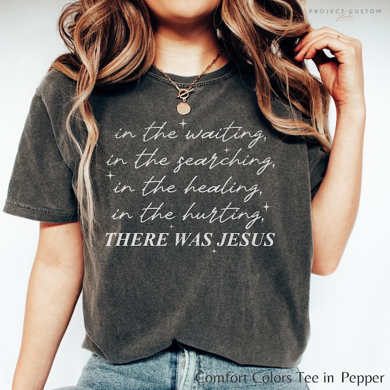 Pu&ograve; includere: Una maglietta grigia scura con il testo "in the waiting, in the searching, in the healing, in the hurting, THERE WAS JESUS" stampato in bianco.