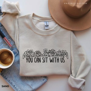 May include: A beige sweatshirt with a black line drawing of the Last Supper and the text "You Can Sit With Us".
