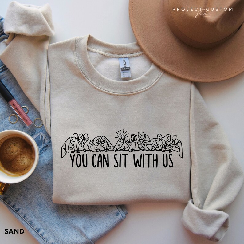 May include: A beige sweatshirt with a black line drawing of the Last Supper and the text "You Can Sit With Us".