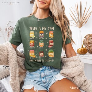 K&ouml;nnte beinhalten: Moosgr&uuml;nes T-Shirt mit dem Text "JESUS IS MY JAM" und Illustrationen von Marmeladengl&auml;sern. Das Shirt enth&auml;lt auch Bibelverse. Das Shirt ist aus einem weichen Material.