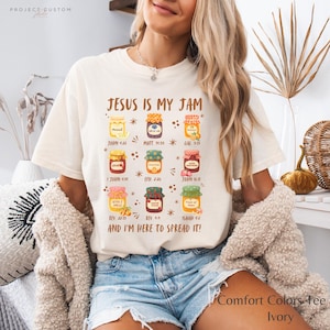 K&ouml;nnte beinhalten: Elfenbeinfarbenes Comfort Colors T-Shirt mit dem Text "JESUS IS MY JAM" und bunten Marmeladenglas-Grafiken. Das Shirt enth&auml;lt Bibelverse wie Johannes 4:26 und Matth&auml;us 19:35. Der Text "AND I'M HERE TO SPREAD IT!" ist ebenfalls aufgedruckt.