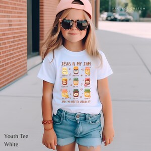 K&ouml;nnte beinhalten: Wei&szlig;es Kinder-T-Shirt mit dem Text "JESUS IS MY JAM" und bunten Marmeladenglas-Grafiken. Das Shirt enth&auml;lt auch den Satz "AND I'M HERE TO SPREAD IT!". Das Model tr&auml;gt blaue Jeansshorts und eine rosa Kappe.