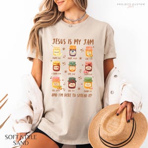K&ouml;nnte beinhalten: Sandfarbenes T-Shirt mit dem Text "JESUS IS MY JAM" und Illustrationen von Marmeladengl&auml;sern. Das Shirt enth&auml;lt auch den Text "AND I'M HERE TO SPREAD IT!" und Bibelverse.
