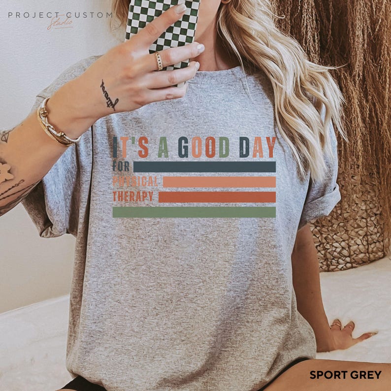 May include: A gray t-shirt with the text "It's a good day for physical therapy" in colorful stripes. The shirt is shown on a model wearing it. The color of the shirt is "Sport Grey".