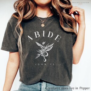 May include: A dark gray t-shirt with the word "ABIDE" in white lettering, a small floral design, and the text "JOHN 15" below. The shirt is made of a soft, comfortable material.