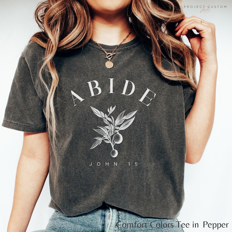 May include: A dark gray t-shirt with the word "ABIDE" in white lettering, a small floral design, and the text "JOHN 15" below. The shirt is made of a soft, comfortable material.