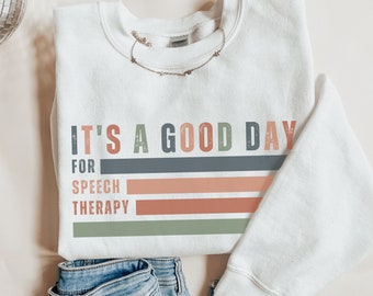 May include: White sweatshirt with the text "It's a good day for speech therapy" in a retro style with colorful stripes.