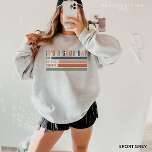 May include: A grey sweatshirt with the text "It's a good day for physical therapy" in a colorful striped design. The sweatshirt is being modeled by a person wearing black shorts.