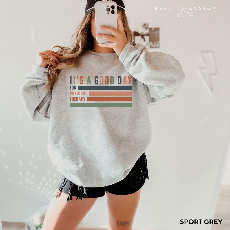 May include: A grey sweatshirt with the text "It's a good day for physical therapy" in a colorful striped design. The sweatshirt is being modeled by a person wearing black shorts.