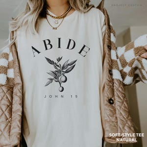 May include: A white t-shirt with the word "ABIDE" printed in black, along with a small black and white illustration of a branch with leaves and fruit. The text "JOHN 15" is printed below the illustration. The shirt is labeled "SOFT-STYLE TEE NATURAL".