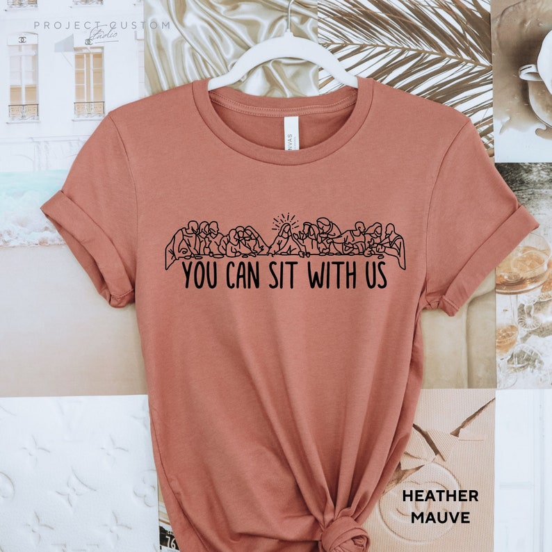 May include: A heather mauve t-shirt with a black graphic of the Last Supper and the text "You Can Sit With Us".