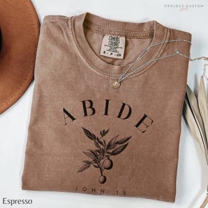 May include: Espresso brown t-shirt with the word "ABIDE" in black lettering, a small branch with leaves and fruit, and the text "JOHN 15" below. The shirt has a distressed look and is made of a soft, comfortable fabric.