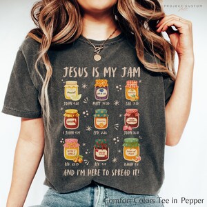 K&ouml;nnte beinhalten: Dunkelgraues Comfort Colors T-Shirt mit dem Text "JESUS IS MY JAM" und bunten Marmeladenglas-Illustrationen. Jedes Glas hat einen Bibelvers-Verweis. Das Shirt sagt auch "AND I'M HERE TO SPREAD IT!"