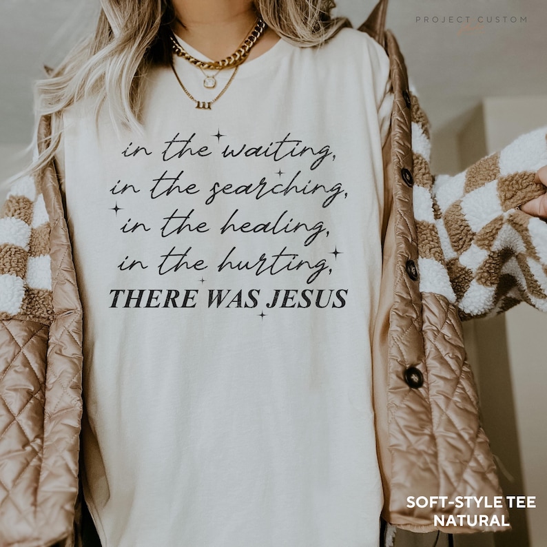 Pu&ograve; includere: Una maglietta bianca con il testo "in the waiting, in the searching, in the healing, in the hurting, THERE WAS JESUS" stampato su di essa. La maglietta &egrave; indossata da una persona con una giacca marrone.