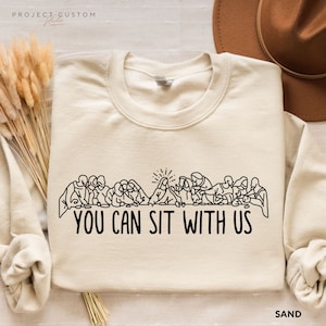 May include: A beige sweatshirt with a black outline of the Last Supper and the text "You Can Sit With Us".