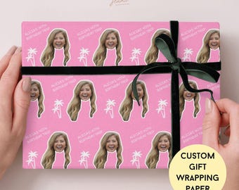 May include: Pink custom gift wrapping paper with a repeating pattern of a woman's face, palm trees, and text. The paper is tied with a black ribbon and bow. The text on the paper reads "ALICIA'S 40TH BIRTHDAY TRIP."