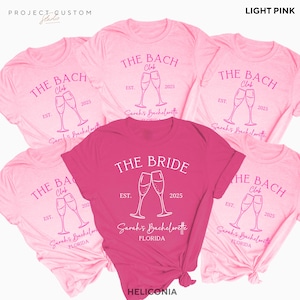 May include: A collection of pink t-shirts with the text "THE BACH Club" and "THE BRIDE" printed on them. The shirts feature a design of two champagne glasses clinking together, with the text "EST. 2025" and "Sarah's Bachelorette FLORIDA".