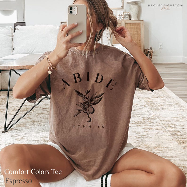 May include: A brown t-shirt with the word "ABIDE" in black lettering, a small floral design, and the text "JOHN 15" underneath. The t-shirt is a comfort colors tee in the color espresso.