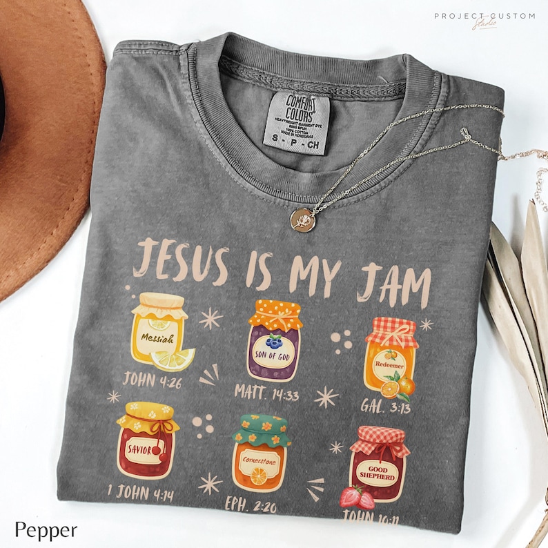 K&ouml;nnte beinhalten: Ein graues Comfort Colors T-Shirt mit dem Schriftzug "JESUS IS MY JAM" und Illustrationen von Marmeladengl&auml;sern. Das Shirt enth&auml;lt Bibelverse wie "JOHN 4:26" und "EPH. 2:20". Eine silberne Halskette mit Anh&auml;nger liegt auf dem Shirt.