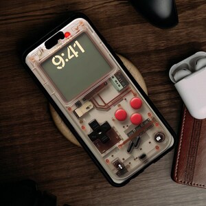 May include: A clear phone case with a retro video game design. The case has a black border and a transparent back that shows the internal components of a video game console. The case has a red and black button design and a digital clock display that reads "9:41".
