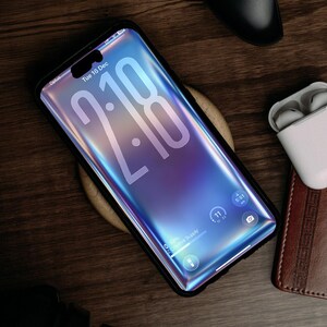 May include: A smartphone with a black case displays the time 2:18 on a gradient blue and purple screen. The phone rests on a wooden surface, with a wireless charger, a controller, and a pair of white earbuds nearby. The phone's screen displays the date, Tuesday, December 16.