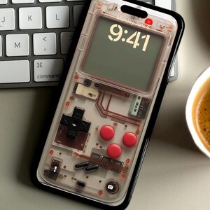 May include: A clear phone case with a retro video game design. The case has a black border and a transparent back that shows the phone's internal components. The case has a digital clock display that shows the time as 9:41. The case also has a red button and a black button.