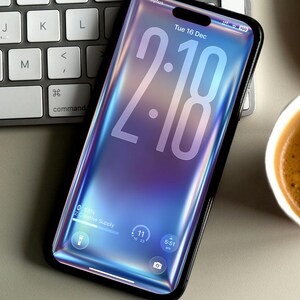 May include: A smartphone with a black frame displays the time 2:18 on a blue and purple gradient background. The screen shows the date, battery life, and other icons. The phone rests on a keyboard.