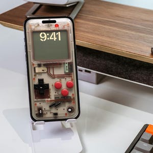 May include: A clear phone case with a retro video game controller design. The case has red buttons and a black and white screen that displays the time and date. The phone is on a white stand. The text on the screen reads "Monday, June 6" and "9:41".