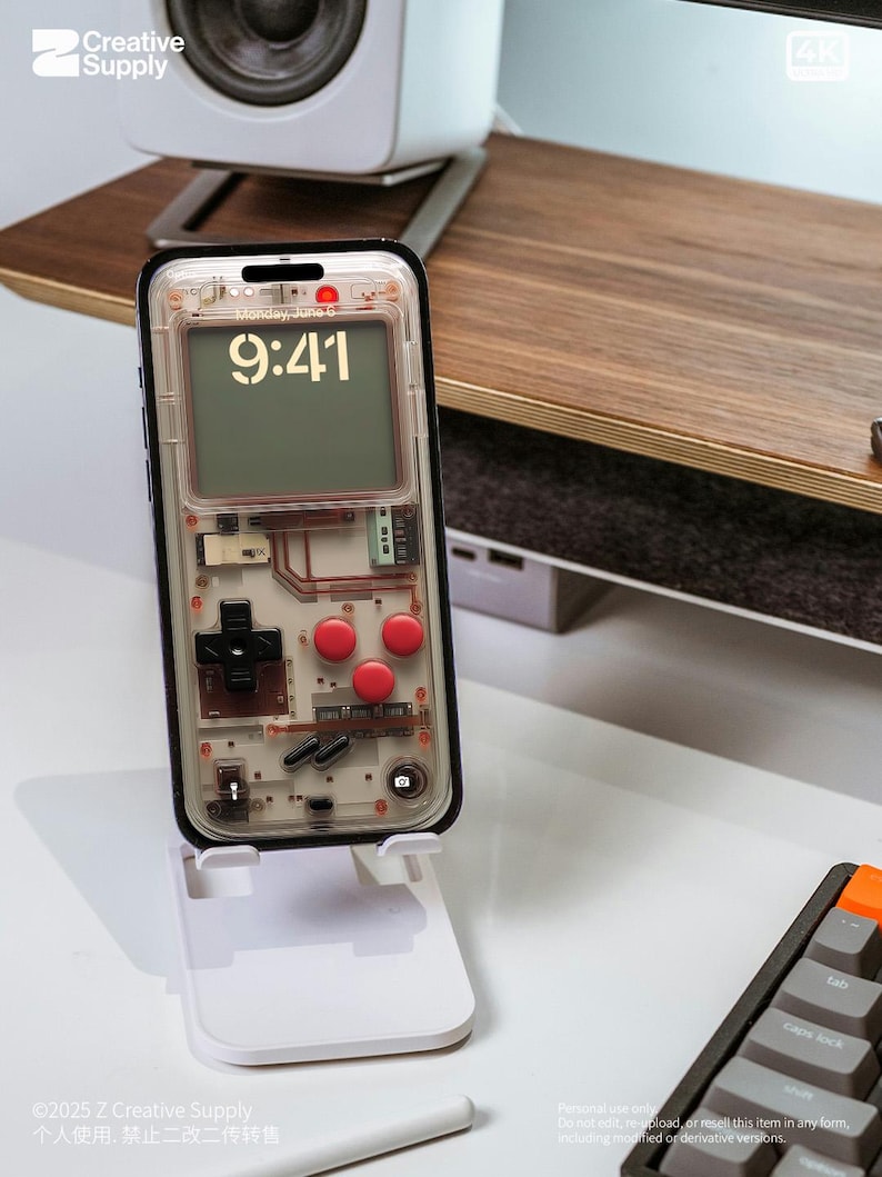May include: A clear phone case with a retro video game controller design. The case has red buttons and a black and white screen that displays the time and date. The phone is on a white stand. The text on the screen reads "Monday, June 6" and "9:41".