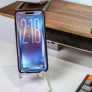 May include: A smartphone with a vibrant blue and purple screen displaying the time 2:18, held by a white phone stand. The phone is on a white desk with a keyboard and a wooden shelf in the background. The phone screen shows the date, Tuesday, December 16.