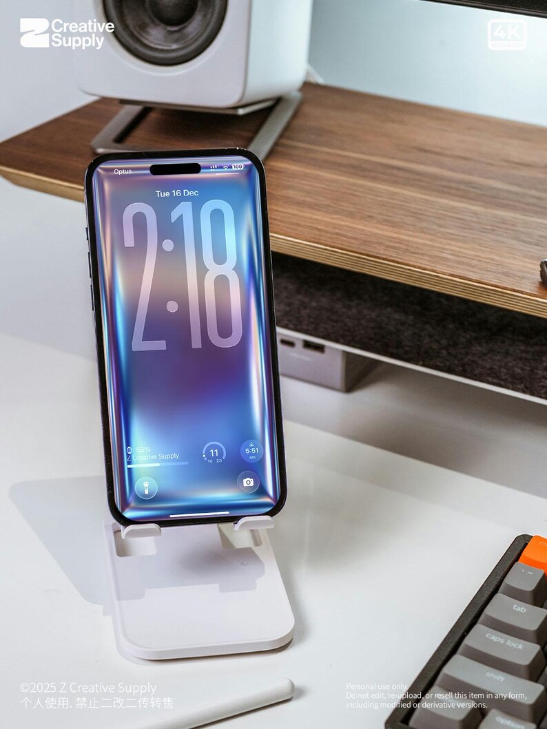 May include: A smartphone with a vibrant blue and purple screen displaying the time 2:18, held by a white phone stand. The phone is on a white desk with a keyboard and a wooden shelf in the background. The phone screen shows the date, Tuesday, December 16.