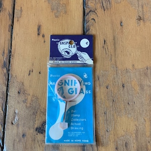 May include: A blue and white card with the text "Magnifying Glass" and "For: Stamp Collectors School Drawing" on it. The card shows a pink magnifying glass with a handle.