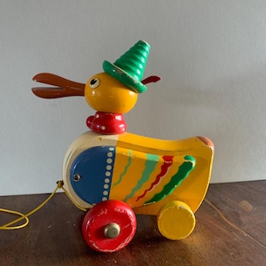 May include: A vintage wooden pull-along duck toy. The duck is painted yellow with a red collar, a green hat, and brown beak. The wheels are red and yellow, and the toy has a yellow pull string. The duck has a blue and white section on its side.