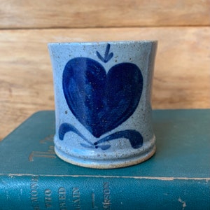 May include: A small, blue and white ceramic mug with a blue heart design. The mug has a speckled glaze and a rounded shape.