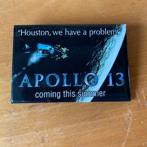 May include: A black and silver button with a picture of a spacecraft and the moon. The text on the button reads "Houston, we have a problem" and "Apollo 13 coming this summer".